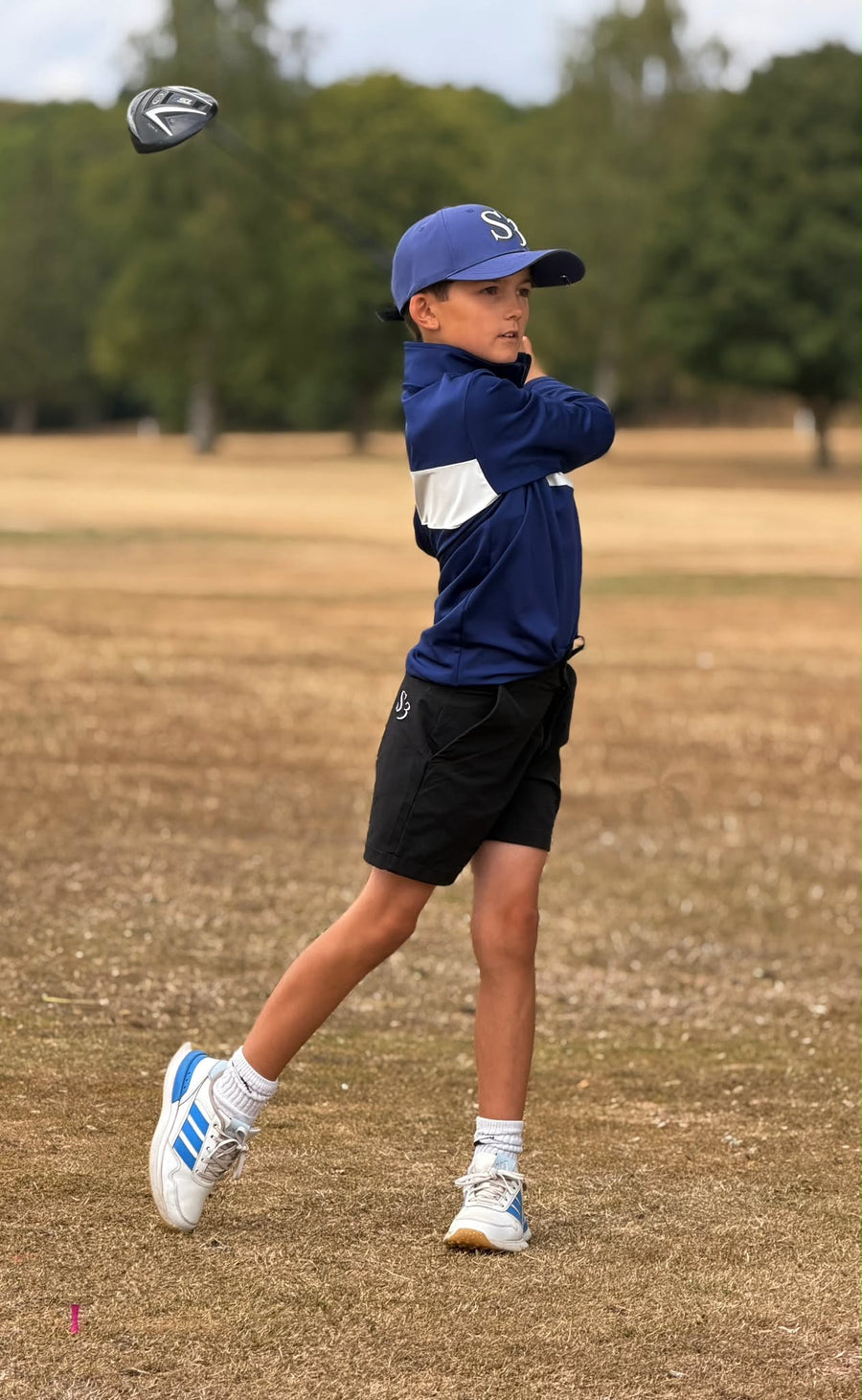 Junior golfer in a navy S3 mid layer, cap and black shorts following through on a driver swing