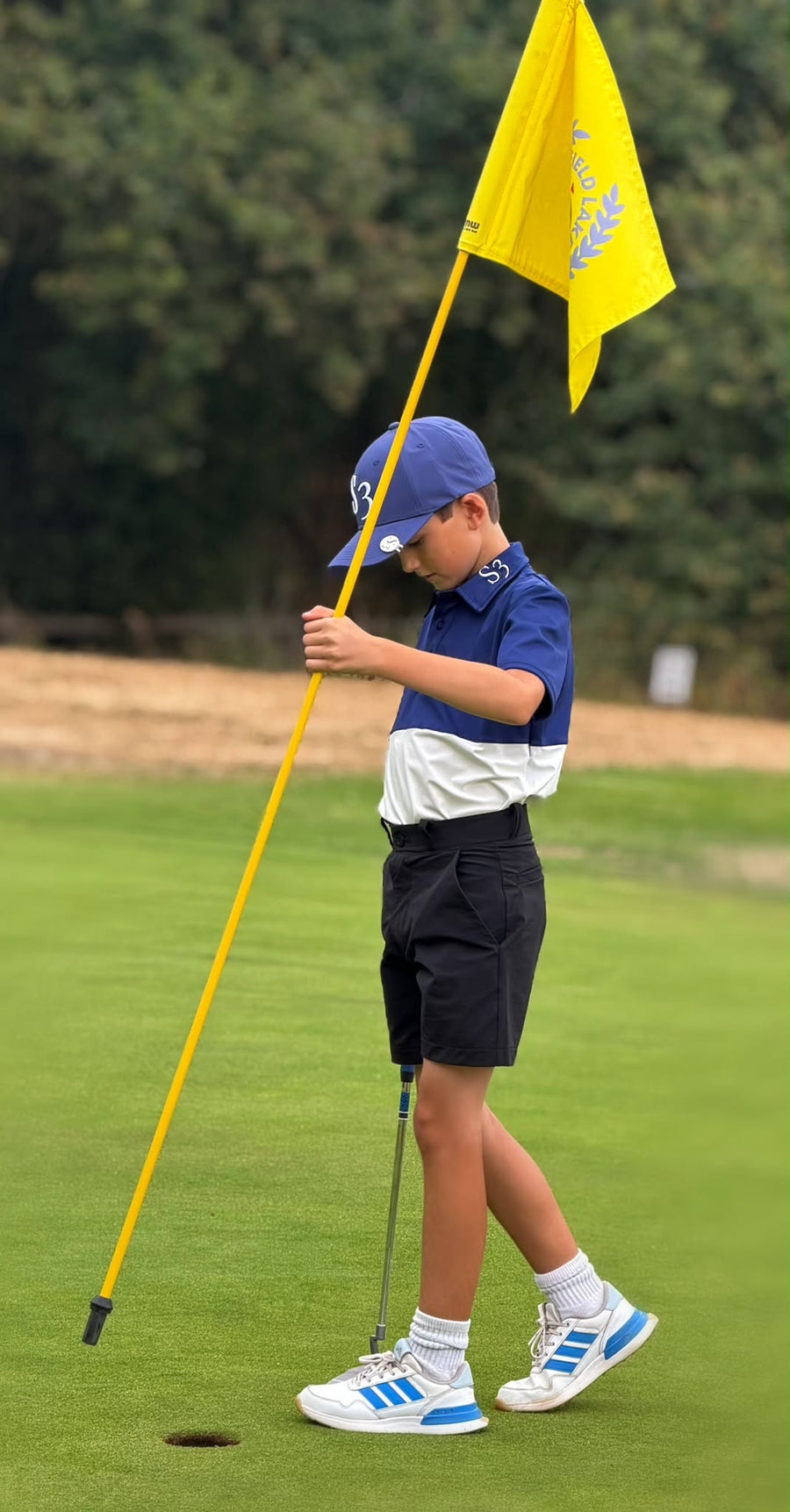 Junior golfer in a navy and white S3 polo and black shorts holding the flagstick on the green