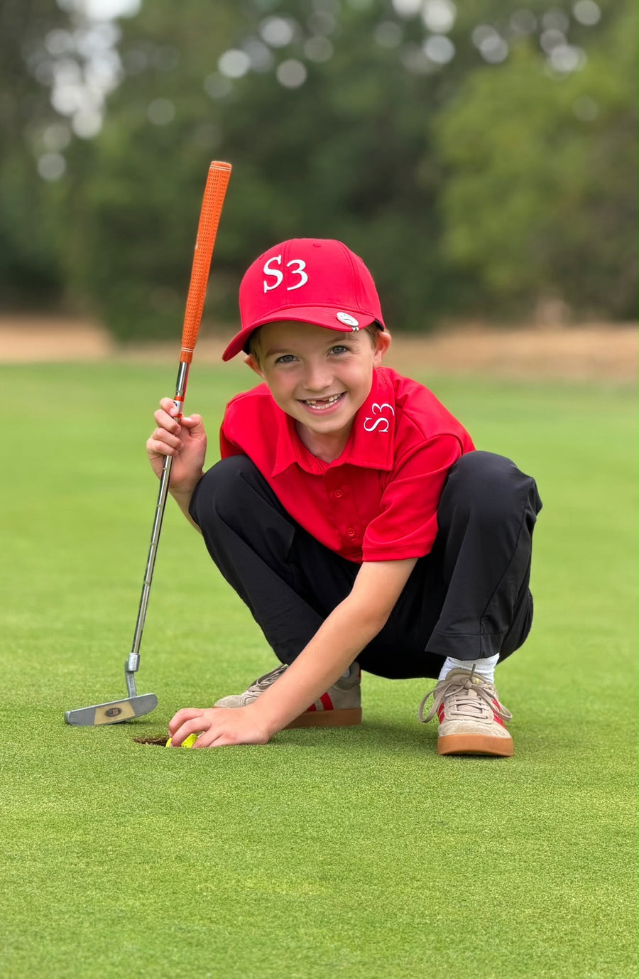 Smiling junior golfer in a red S3 polo and cap crouching by the hole with a putter