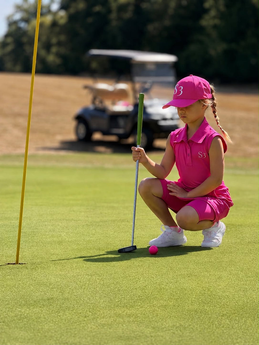 Junior golfer in a pink S3 sleeveless polo and cap crouching to line up a putt