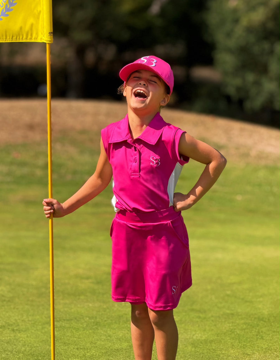 Laughing junior golfer in a pink S3 polo, skort and cap holding the flagstick