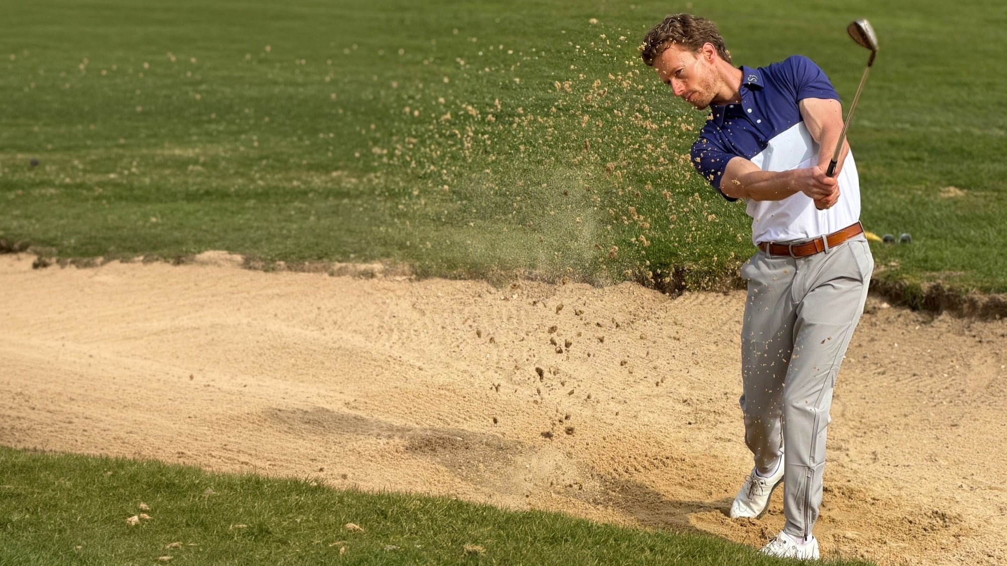 Golfer in an S3 polo playing out of a greenside bunker, sand spraying